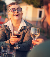 Happy beautiful mature woman in glasses 60 years old sitting in a cafe with her girlfriend drinking wine in a street cafe