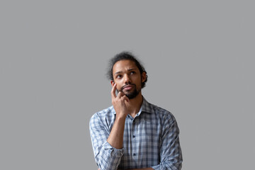 Unhappy thoughtful African American man looking up, standing isolated on grey studio wall background, young male lost in thoughts, thinking about personal problems, making difficult decision