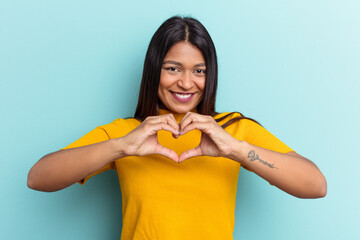Young Venezuelan woman isolated on blue background smiling and showing a heart shape with hands.