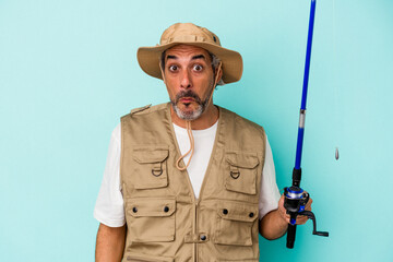Middle age caucasian fisherman holding rod isolated on blue background  shrugs shoulders and open eyes confused.