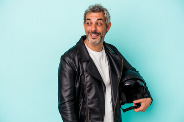 Middle age biker caucasian man holding helmet isolated on blue background  looks aside smiling,...