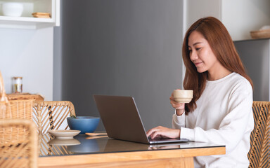 A young woman freelancer drinking coffee while using laptop computer for working or studying online at home