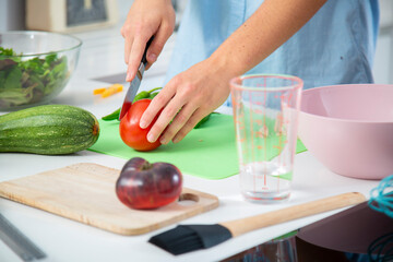 manos de chico joven picando tomate en una tabla de cortar verde con tabla de madera, `pimientos, calabacin, vaso de cristal para harina, bol rosa close-up - obrazy, fototapety, plakaty