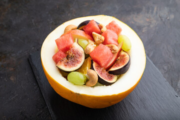 Vegetarian fruit salad of watermelon, grapes, figs, pear, orange, cashew  on black concrete background. Side view, close up.