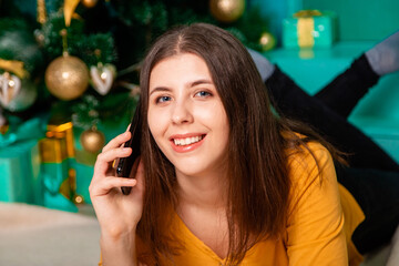 A young woman lies on a fur carpet with a smartphone in her hands on the background of a Christmas tree.