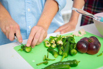 close-up manos de chico joven corta pimientos verdes en una tabla de cortar verde con su pareja en una cocina blanca en la que hay mas verduras como tomate, calabacin y lechuga o un bol rosa 