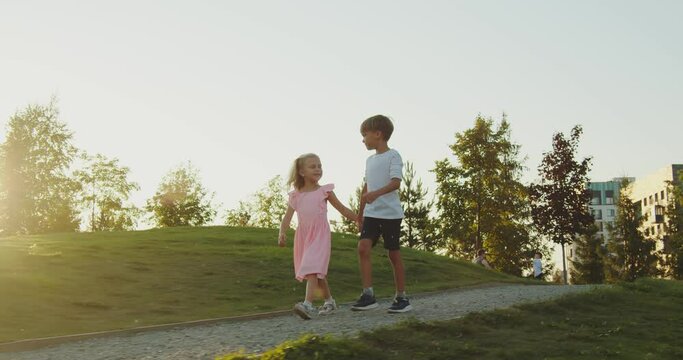 Children walk in the park hand in hand. This is a brother and sister walking along the path in the park, waving their arms. Around them there is green grass and trees, the sun is shining brightly
