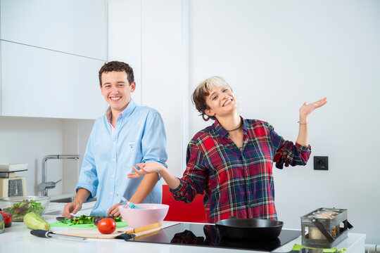 pareja joven sonriente cocina pimiento verde con una sarten de hierro en una cocina blanca con verduras, con placa de induccion, utilizan un bol rosa y usan herramientas de cocina