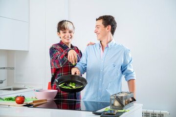 pareja joven sonriente cocinan con una sarten de hierro verduras, pimiento verde, tomate y...