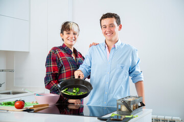 pareja joven sonriente cocinan con una sarten de hierro verduras, pimiento verde, tomate y calabacin, en una cocina blanca con placa de induccion, utilizan un bol rosa y  herramientas de cocina