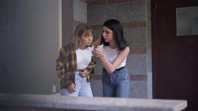 Portrait of angry teenage girl threatening classmate showing social media on smartphone. Aggressive Caucasian girl arguing with nerd groupmate outdoors for online bullying