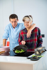 mujer joven sonriente cocina con su pareja pimiento verde, tomate y calabacin, en una cocina blanca...