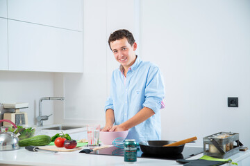 hombre joven sonriente hace una masa en un bol rosa, en una cocina blanca con placa de induccion, ...