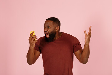 Angry African-American man in brown t-shirt screams holding modern mobile phone on light pink background in studio close view - 459452385
