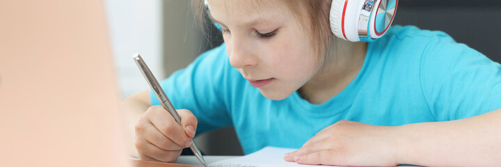 Little girl in headphones sitting at table in front of laptop and drawing