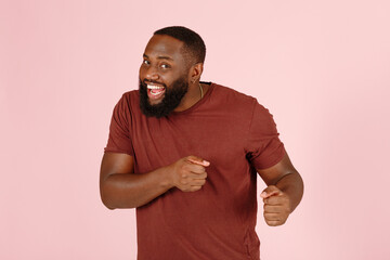 Positive bearded African-American guy actor in brown t-shirt dances standing on light pink background in studio close view - 459452764