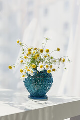 Daisies vase blue on a white table. Beautiful bright summer still life with wild flowers. A gift for a birthday, anniversary, mother. Bright sunlight with beautiful shadows. Vertical postcard design