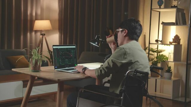 Asian Man Trader Investor Sitting In A Wheelchair And Drink Coffee While Using Laptop Computer For Cryptocurrency Financial Market Analysis, Trading Data Index Chart Graph On Laptop Screen. Back View
