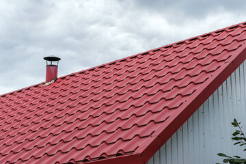 A roof made of red metal tiles with a chimney against a cloudy sky.