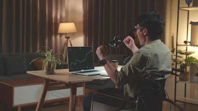 Asian Man Trader Investor Sitting In A Wheelchair Celebrating While Using Laptop Computer For Cryptocurrency Financial Market Analysis, Trading Data Index Chart Graph On Laptop Screen. Back View
