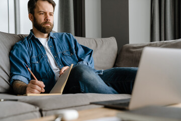 Bearded man writing down notes while working with laptop on sofa