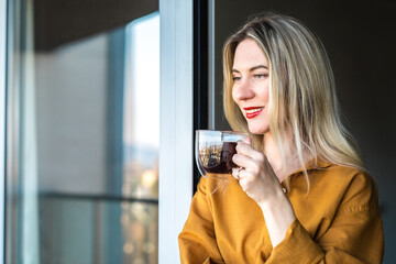 Beautiful smiling Businesswoman in glasses standing near the window and drinking tea cup. Female pretty face, work from home, social distancing, alone at home in office. close up front portrait