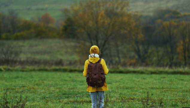 Back view female tourist with backpack standing alone at green lawn among hills. Cold weather, misty autumn nature view