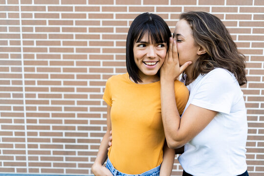 Woman whispering in girlfriend ear in front of brick wall