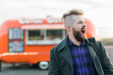 Bearded man in the street yawns with opened mouth stands at a food truck background. Sleepy people and lazy concept.