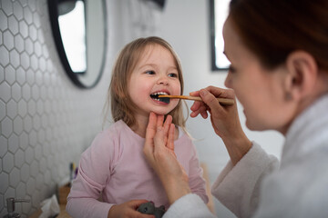 Mother with small daughter brushing teeth indoors in bathroom in the evening or morning.