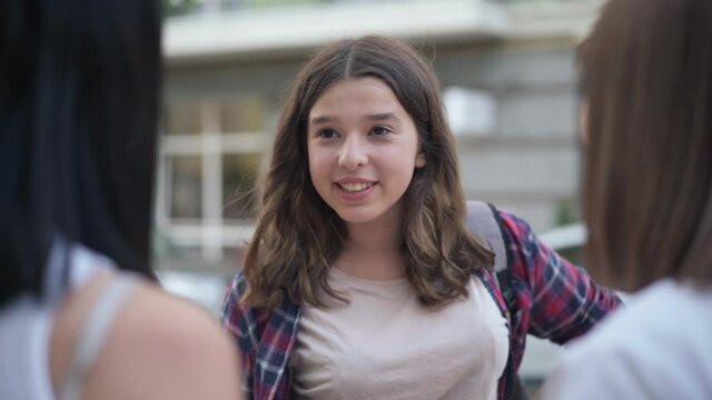 Portrait of charming beautiful teenage girl meeting friends on city street talking smiling. Positive Caucasian teenager chatting with girlfriends sharing rumors outdoors. Lifestyle and joy concept