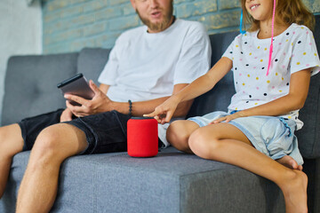 Father and daughter testing talking smart wireless speaker, controlling home devices with a voice commands, sitting on sofa at home, Concept of smart home