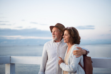Happy senior couple hugging outdoors on pier by sea, looking at view.