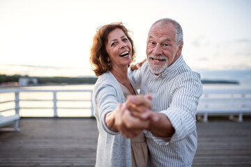 Happy senior couple dancing outdoors on pier by sea, looking at camera.