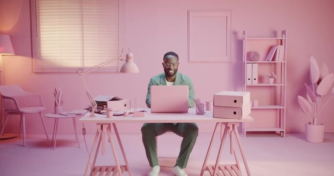 Young happy black business man sits at table with laptop working in pink office. Gets great news or reading email or enjoying social networking at monochrome rose pastel design room studio. Monocolor