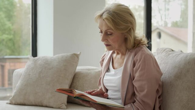 Elderly woman resting at living room. She is reading novel 