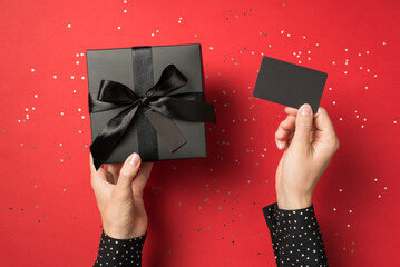 First person view overhead photo of black giftbox with ribbon wrapped as bow and business card in hands and confetti around isolated on the red background - Powered by Adobe
