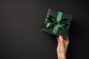 First person top view photo of hand holding black giftbox with green satin ribbon bow on isolated black background with copyspace