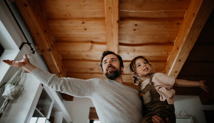 Low angle view of mature father with small daughter standing indoors at home, holding and hugging.