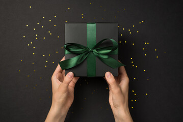 First person top view photo of hands giving black gift box with green ribbon bow and golden...