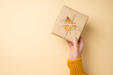First person top view photo of hand in orange sweater giving craft paper giftbox with twine bow and yellow autumn leaf on isolated beige background with empty space
