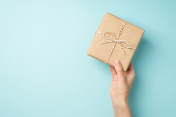 First person top view photo of female hand holding stylish craft paper giftbox with twine bow on isolated pastel blue background with copyspace