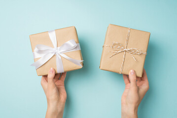 First person top view photo of female hands holding two craft paper gift boxes with twine bow and white satin ribbon bow on isolated pastel blue background
