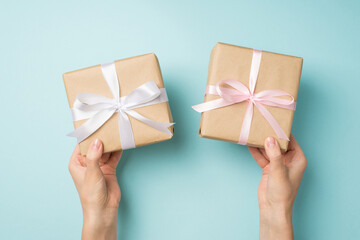First person top view photo of hands holding two craft paper gift boxes with pink and white satin ribbon bows on isolated pastel blue background