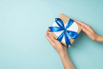 First person top view photo of hands holding craft paper giftbox with blue ribbon bow and white paper heart on isolated pastel blue background with copyspace