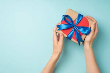 First person top view photo of female hands holding craft paper giftbox with blue ribbon bow and red paper heart on isolated pastel blue background with copyspace