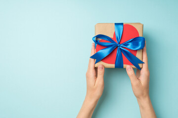 First person top view photo of hands giving craft paper giftbox with blue ribbon bow and red paper heart on isolated pastel blue background with empty space