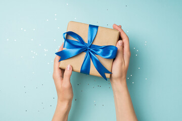First person top view photo of female hands holding craft paper giftbox with blue ribbon bow over shiny confetti on isolated pastel blue background