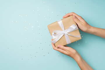 First person top view photo of hands demonstrating craft paper gift box with white satin ribbon bow over sequins on isolated pastel blue background with copyspace