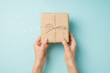 First person top view photo of hands holding craft paper gift box with twine bow over sequins on isolated pastel blue background